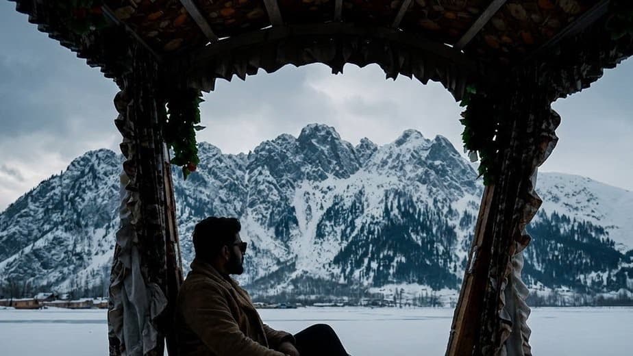 A man sits in the shade of a carved wooden boat canopy, looking out across a frozen lake at snow covered mountains under heavy cloud.